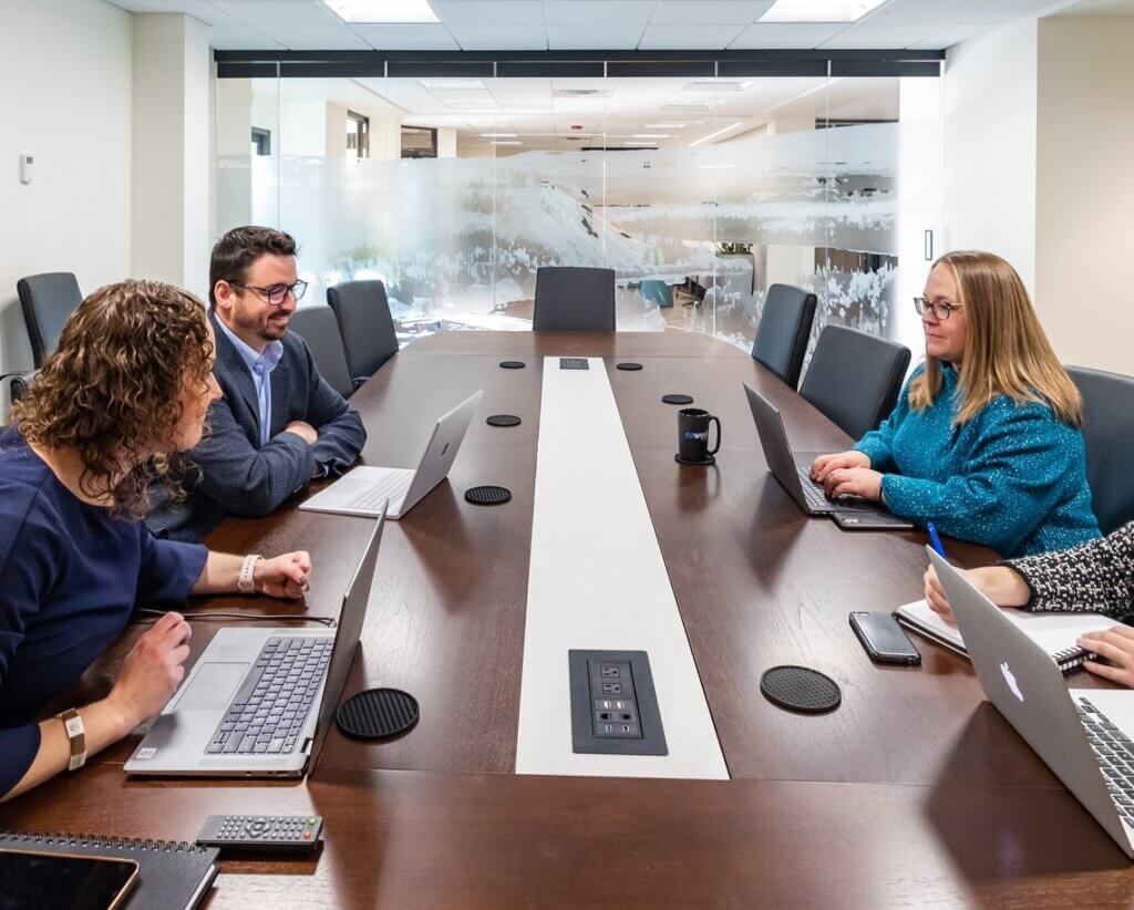 A meeting in a conference room at our coworking space, Cowork by RSD in Anchorage, Alaska. There is a woman and man sitting on the left and a woman on the right of a long, dark conference room table. All are using laptops. Glass with an etched mountain scene is in the background.