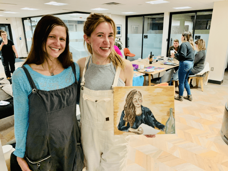 Two women with an arm around each other, with the woman on the right holding a self-portrait. Other art workshop participants are in the background.